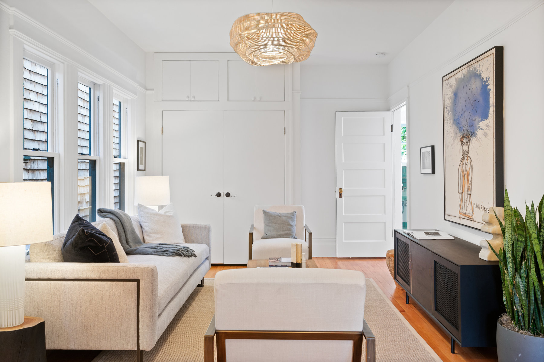 A centered shot of the living room. Large windows on the left side and there is a doorway to kitchen in back right corner of room.