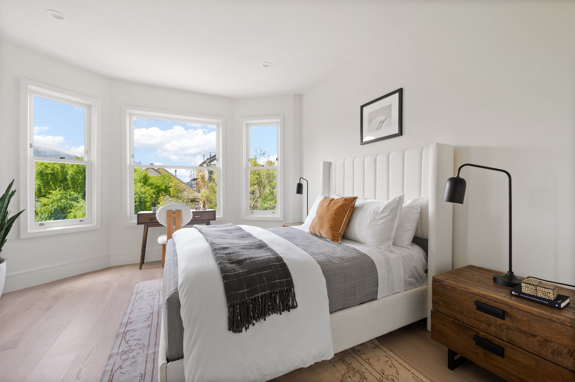 Guest bedroom has large bay windows with small desk under. There is a queen bed with two bedside tables.