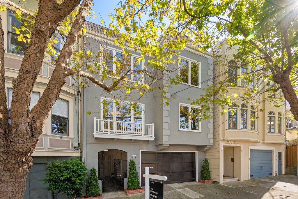 Gray two-story townhouse with white trim, a balcony, and garage, situated between similar homes on a tree-lined street in daylight.