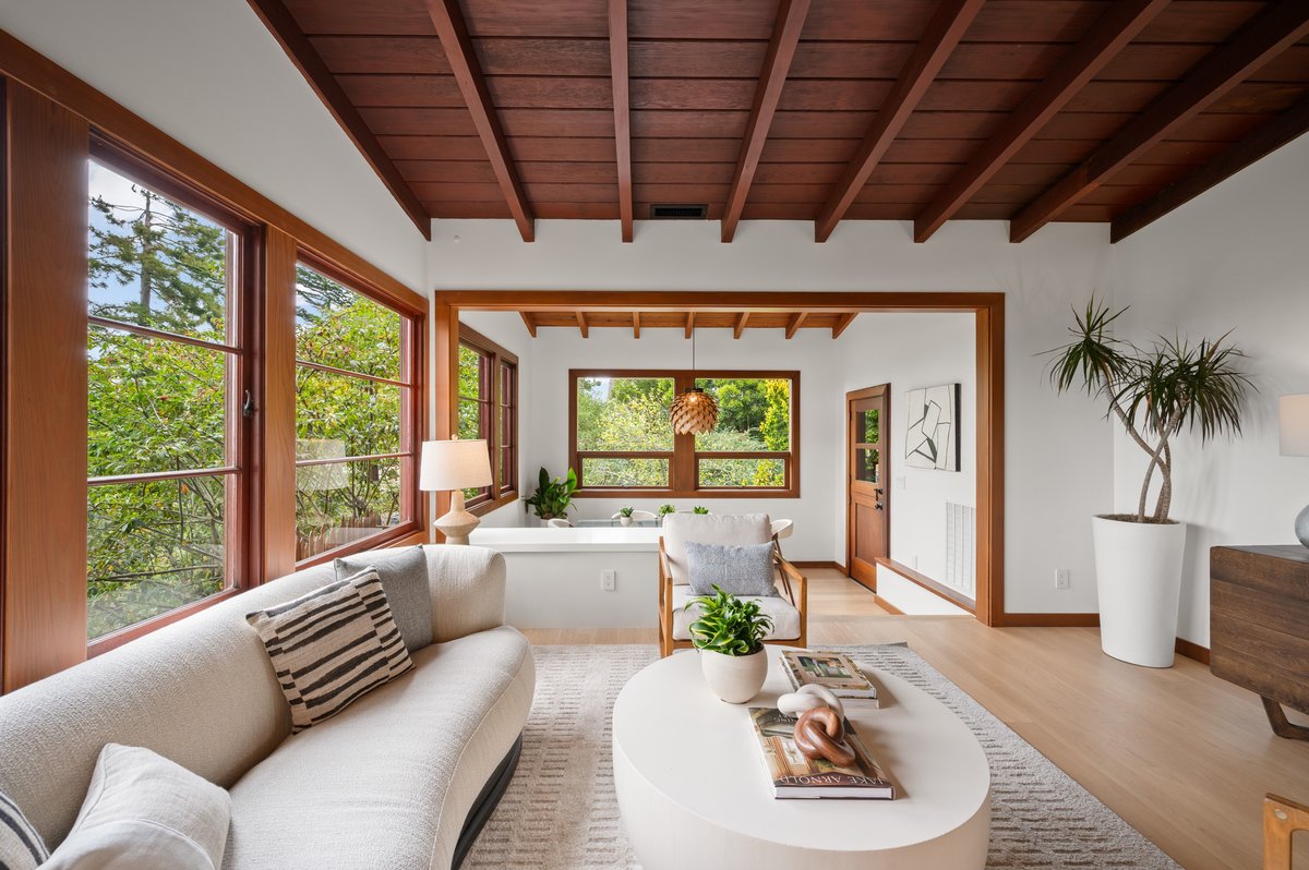 Modern living room with wood ceiling, large windows, white walls, light-colored furniture, plants, and a round coffee table with magazines and decor. Modern living room with wood ceiling, large windows, white walls, light-colored furniture, plants, and a round coffee table with magazines and decor.