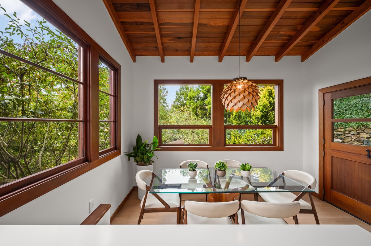 A dining area with a glass table, four white chairs, potted plants, large windows, wooden ceiling, and a wooden door with glass panels, overlooking greenery outside. A dining area with a glass table, four white chairs, potted plants, large windows, wooden ceiling, and a wooden door with glass panels, overlooking greenery outside.