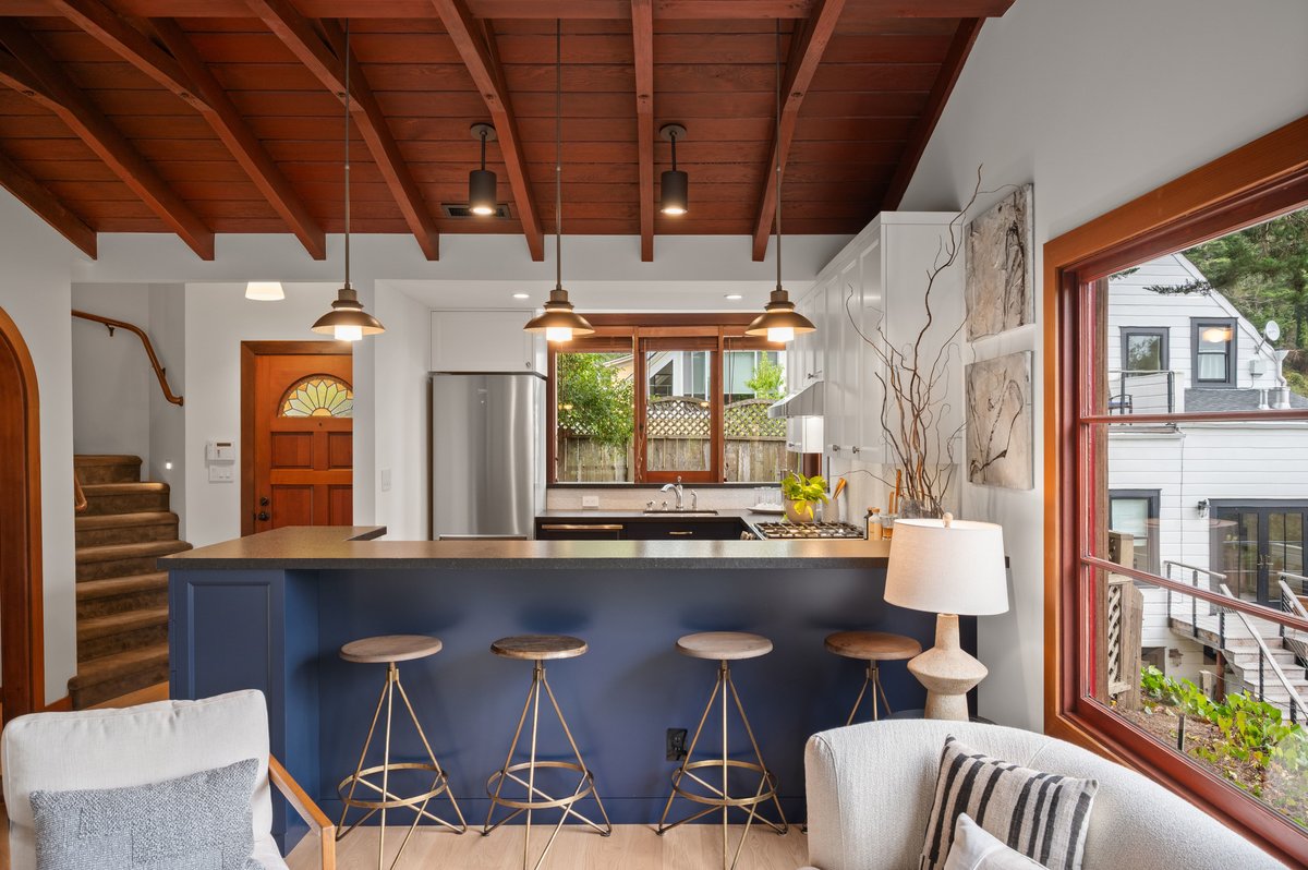 Modern kitchen with blue island, three stools, pendant lights, exposed wood ceiling, large window, and adjacent living area with lamp and armchair. Modern kitchen with blue island, three stools, pendant lights, exposed wood ceiling, large window, and adjacent living area with lamp and armchair.