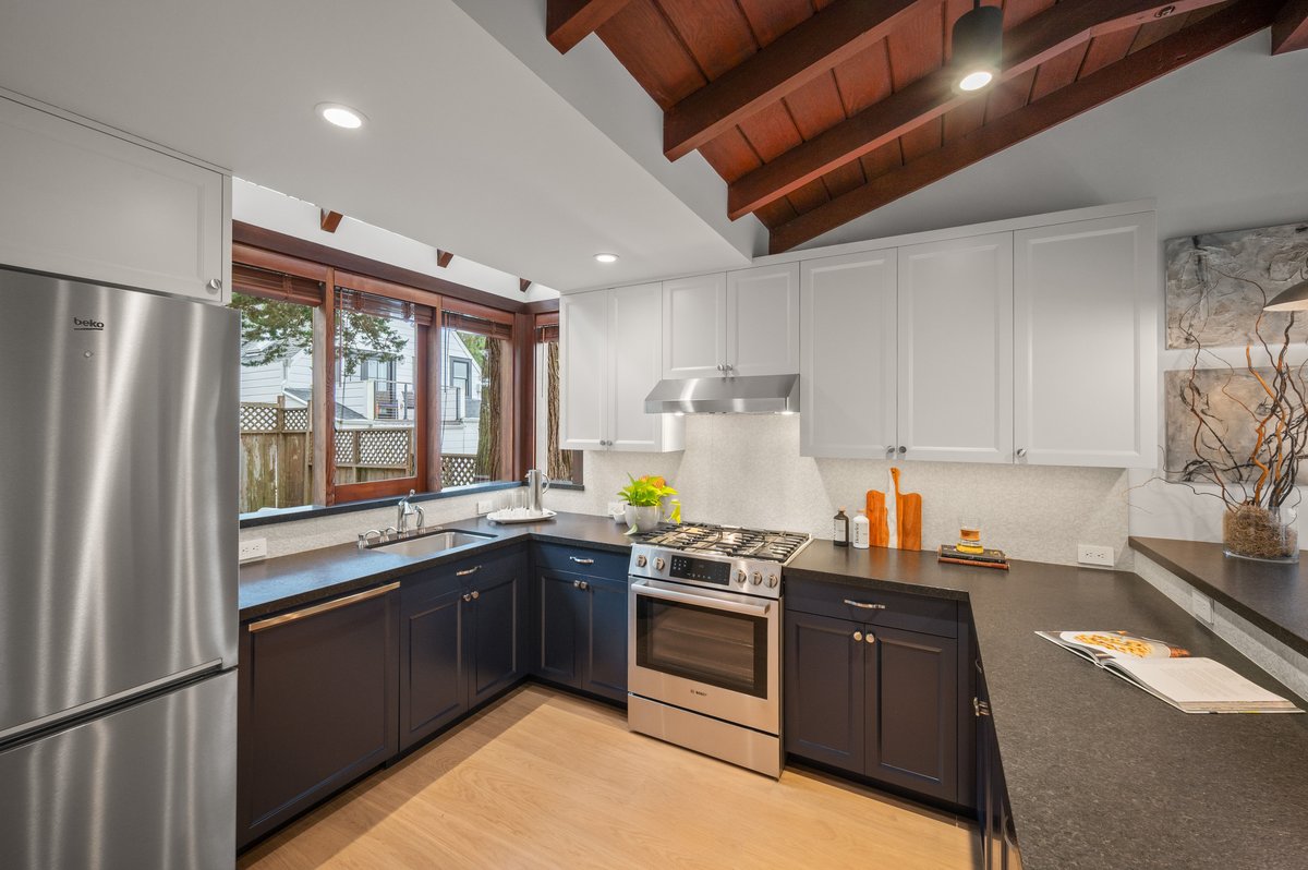 Modern kitchen with white upper cabinets, dark lower cabinets, stainless steel appliances, wood ceiling beams, and large windows overlooking a fenced backyard. Modern kitchen with white upper cabinets, dark lower cabinets, stainless steel appliances, wood ceiling beams, and large windows overlooking a fenced backyard.