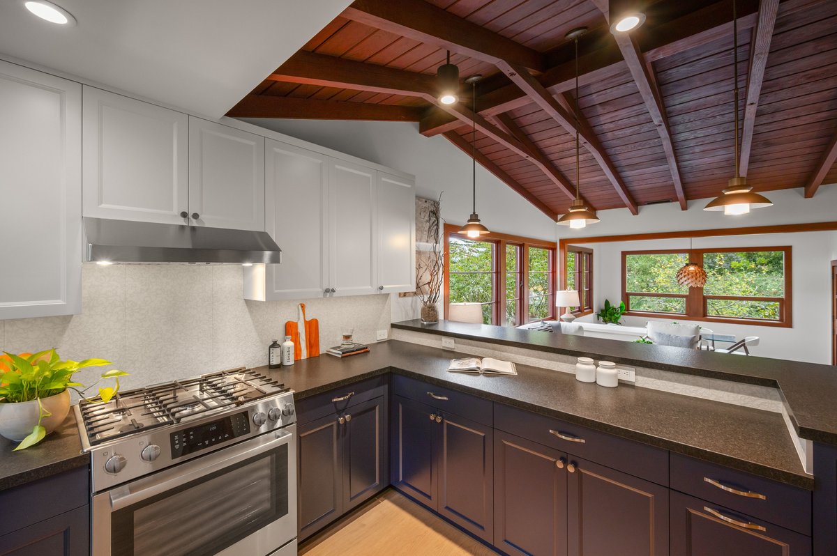 Modern kitchen with dark countertops, navy and white cabinets, stainless steel stove, and wood ceiling, open to a bright living area with large windows and greenery outside. Modern kitchen with dark countertops, navy and white cabinets, stainless steel stove, and wood ceiling, open to a bright living area with large windows and greenery outside.
