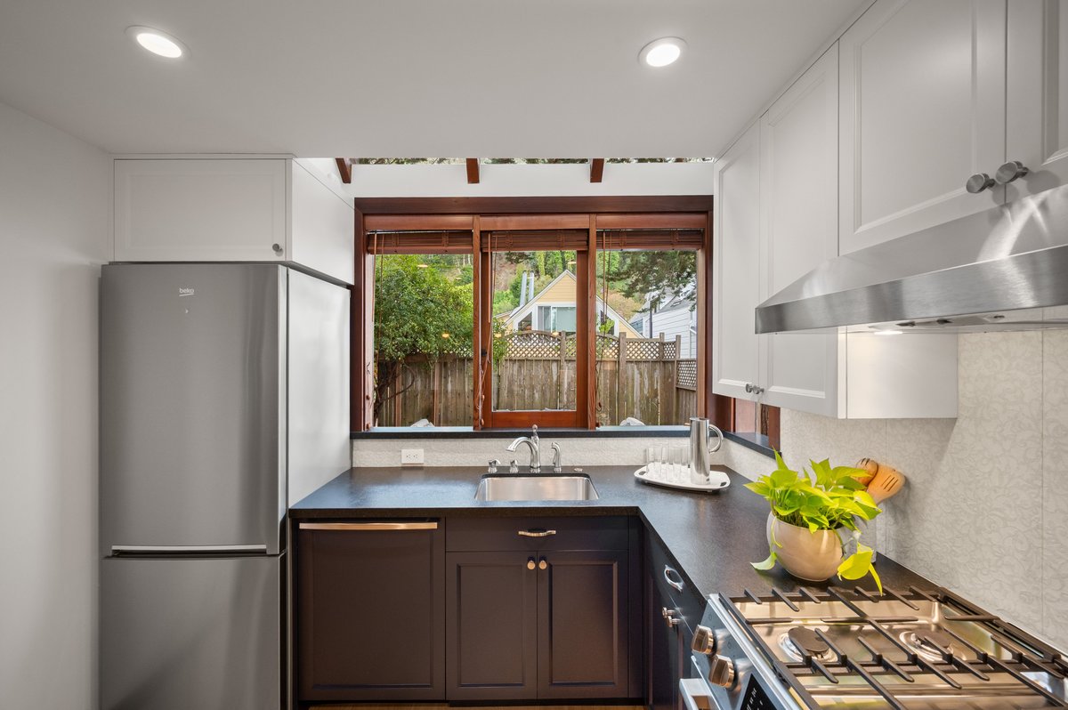 Modern kitchen with white cabinets, stainless steel appliances, a sink under a large window, black countertops, and a potted plant next to a gas stove with range hood. Trees visible outside. Modern kitchen with white cabinets, stainless steel appliances, a sink under a large window, black countertops, and a potted plant next to a gas stove with range hood. Trees visible outside.