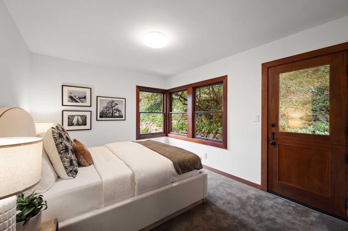 A bedroom with a bed, framed photos on the wall, a side table with a lamp, a corner window showing greenery, and a wooden door leading outside. A bedroom with a bed, framed photos on the wall, a side table with a lamp, a corner window showing greenery, and a wooden door leading outside.