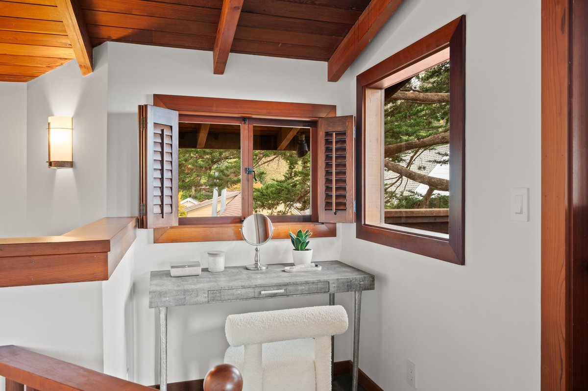 A small vanity with a stool sits by two windows with wooden shutters in a room with white walls and wood trim. A small vanity with a stool sits by two windows with wooden shutters in a room with white walls and wood trim.