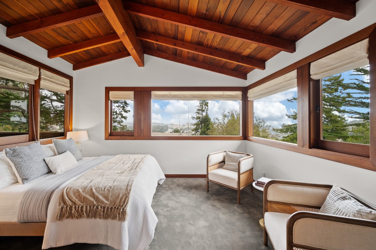 Bedroom with a wooden beamed ceiling, large corner windows, a made bed, and two armchairs, offering a view of trees and the sky outside. Bedroom with a wooden beamed ceiling, large corner windows, a made bed, and two armchairs, offering a view of trees and the sky outside.