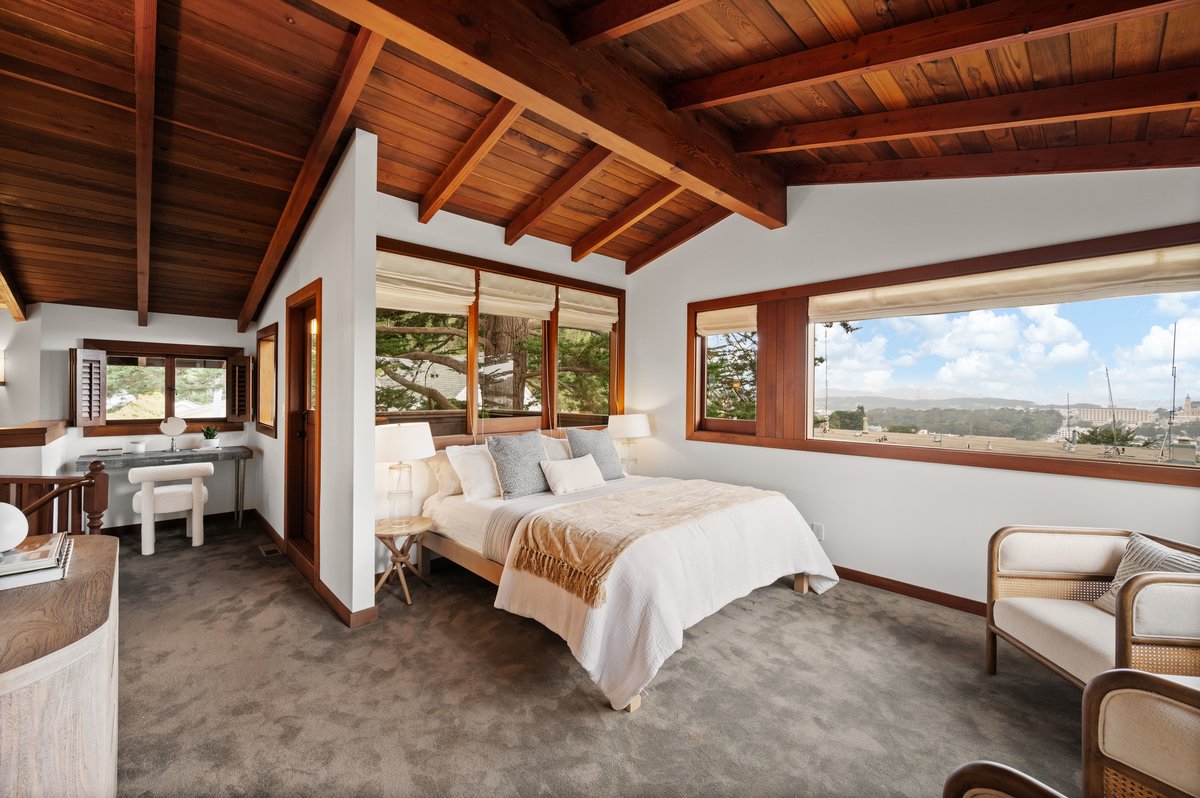Bedroom with wood ceiling, large windows with outdoor views, a bed with white bedding, two chairs, and a desk area in the corner. Natural light fills the carpeted room. Bedroom with wood ceiling, large windows with outdoor views, a bed with white bedding, two chairs, and a desk area in the corner. Natural light fills the carpeted room.