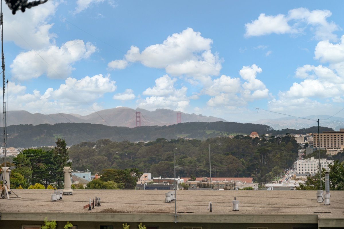 View of the Golden Gate Bridge in the distance, partially obscured by fog, with trees, buildings, and a rooftop in the foreground under a partly cloudy sky. View of the Golden Gate Bridge in the distance, partially obscured by fog, with trees, buildings, and a rooftop in the foreground under a partly cloudy sky.
