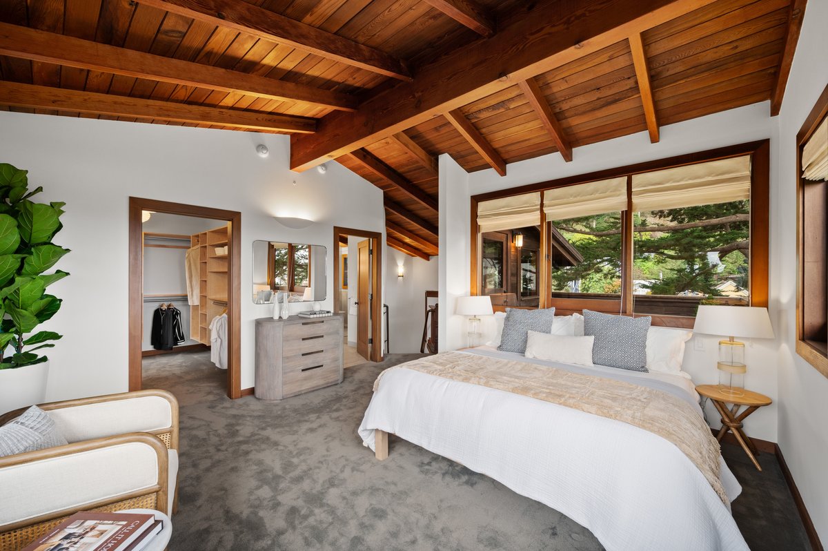 A modern bedroom with a wooden ceiling, large windows, a neatly made bed, nightstands with lamps, a potted plant, and an open doorway leading to a walk-in closet and bathroom. A modern bedroom with a wooden ceiling, large windows, a neatly made bed, nightstands with lamps, a potted plant, and an open doorway leading to a walk-in closet and bathroom.