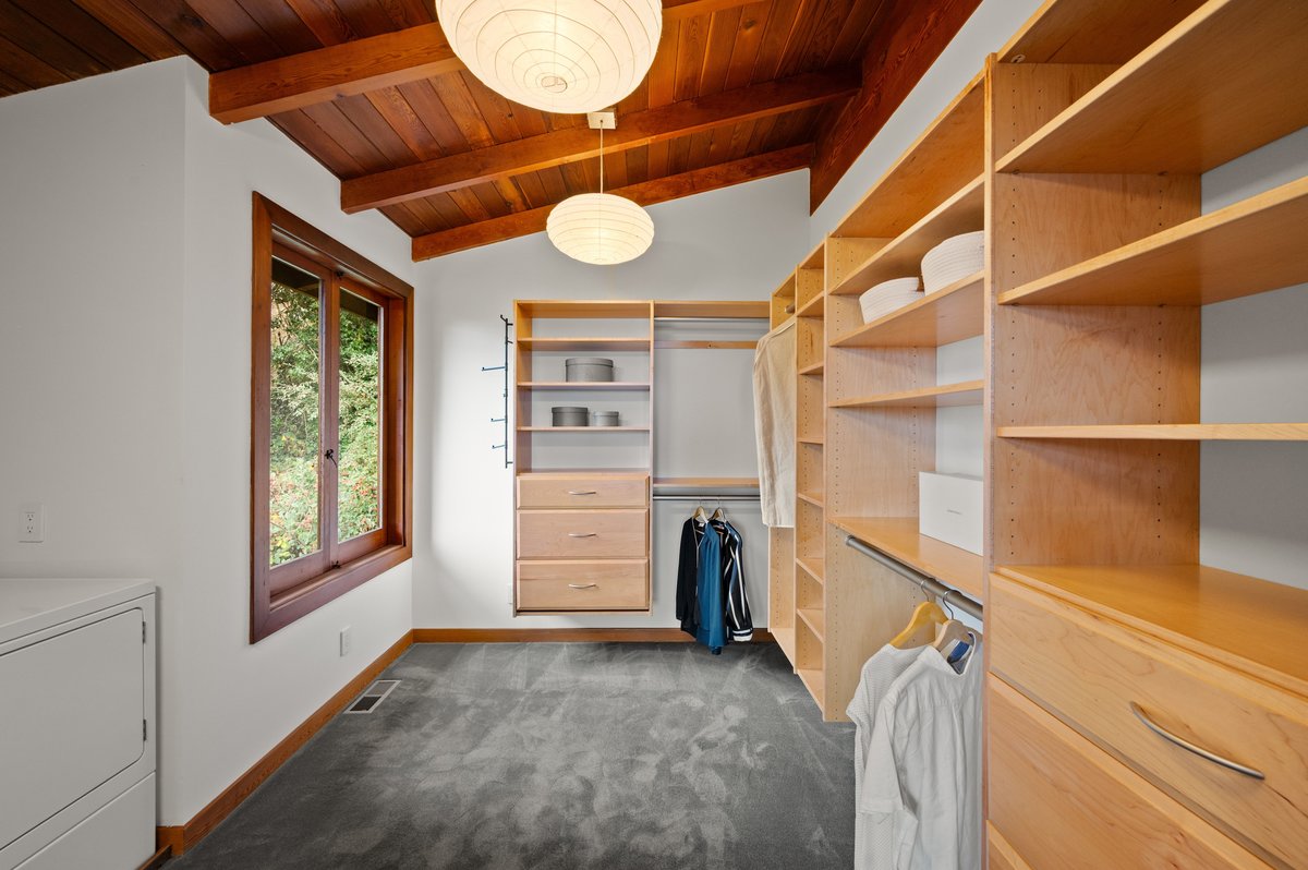 Walk-in closet with wooden shelves and drawers, hanging shirts and jackets, a window with a tree view, gray carpet, and two round ceiling lights. Walk-in closet with wooden shelves and drawers, hanging shirts and jackets, a window with a tree view, gray carpet, and two round ceiling lights.
