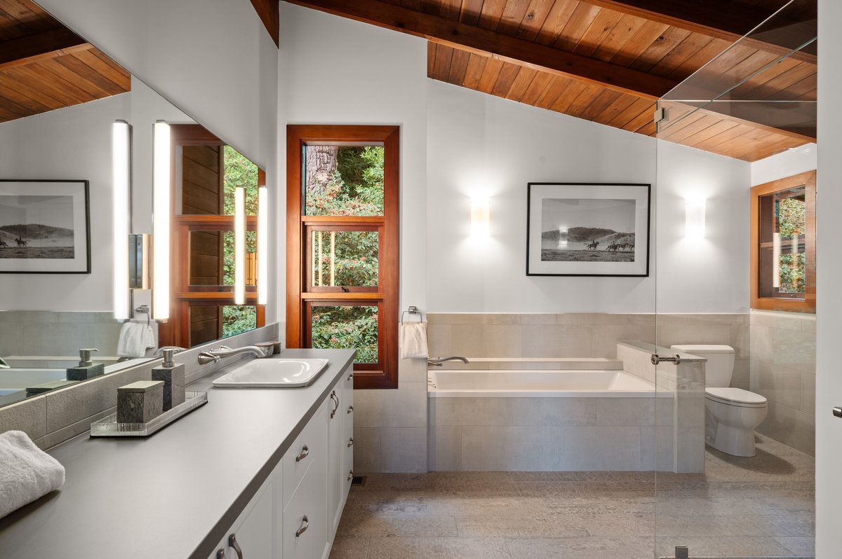 Modern bathroom with a wood ceiling, large mirror, bathtub, toilet, white cabinets, and a window letting in natural light. Modern bathroom with a wood ceiling, large mirror, bathtub, toilet, white cabinets, and a window letting in natural light.