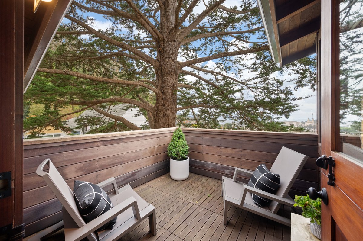 Small wooden balcony with two lounge chairs, black and white pillows, a potted plant, and a large tree in the background under a partly cloudy sky. Small wooden balcony with two lounge chairs, black and white pillows, a potted plant, and a large tree in the background under a partly cloudy sky.