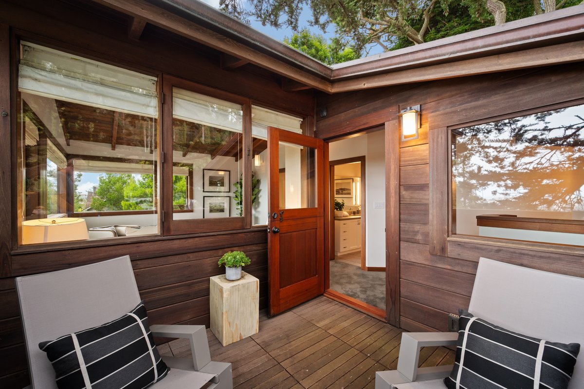 A small wooden patio with two chairs and a potted plant leads to an open door, revealing a cozy interior with warm lighting and modern decor. A small wooden patio with two chairs and a potted plant leads to an open door, revealing a cozy interior with warm lighting and modern decor.