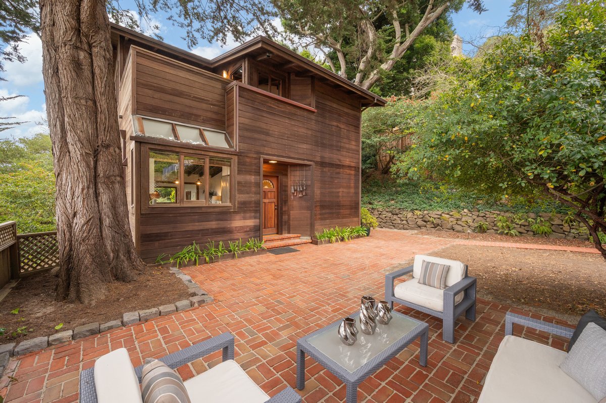 Two-story wooden house with large windows, surrounded by trees. Brick patio features outdoor seating and a coffee table with a metallic tea set. Stone retaining wall visible in the background. Two-story wooden house with large windows, surrounded by trees. Brick patio features outdoor seating and a coffee table with a metallic tea set. Stone retaining wall visible in the background.