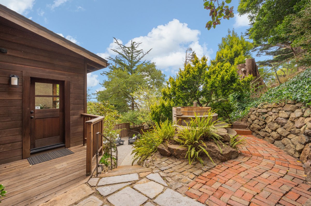 Wooden house with deck, curved brick walkway, stone wall, plants, trees, and a round hot tub in a landscaped backyard on a sunny day. Wooden house with deck, curved brick walkway, stone wall, plants, trees, and a round hot tub in a landscaped backyard on a sunny day.