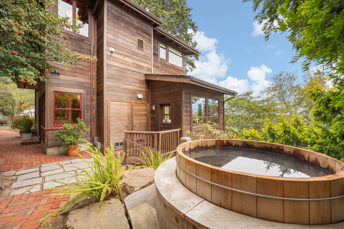 A two-story wooden house with large windows sits beside a circular outdoor hot tub, surrounded by greenery and brick paving under a blue sky with clouds. A two-story wooden house with large windows sits beside a circular outdoor hot tub, surrounded by greenery and brick paving under a blue sky with clouds.