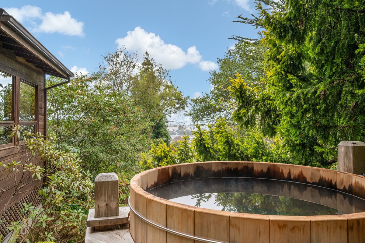 A round wooden hot tub sits on a deck surrounded by trees, with a partial view of distant hills under a partly cloudy sky. A round wooden hot tub sits on a deck surrounded by trees, with a partial view of distant hills under a partly cloudy sky.