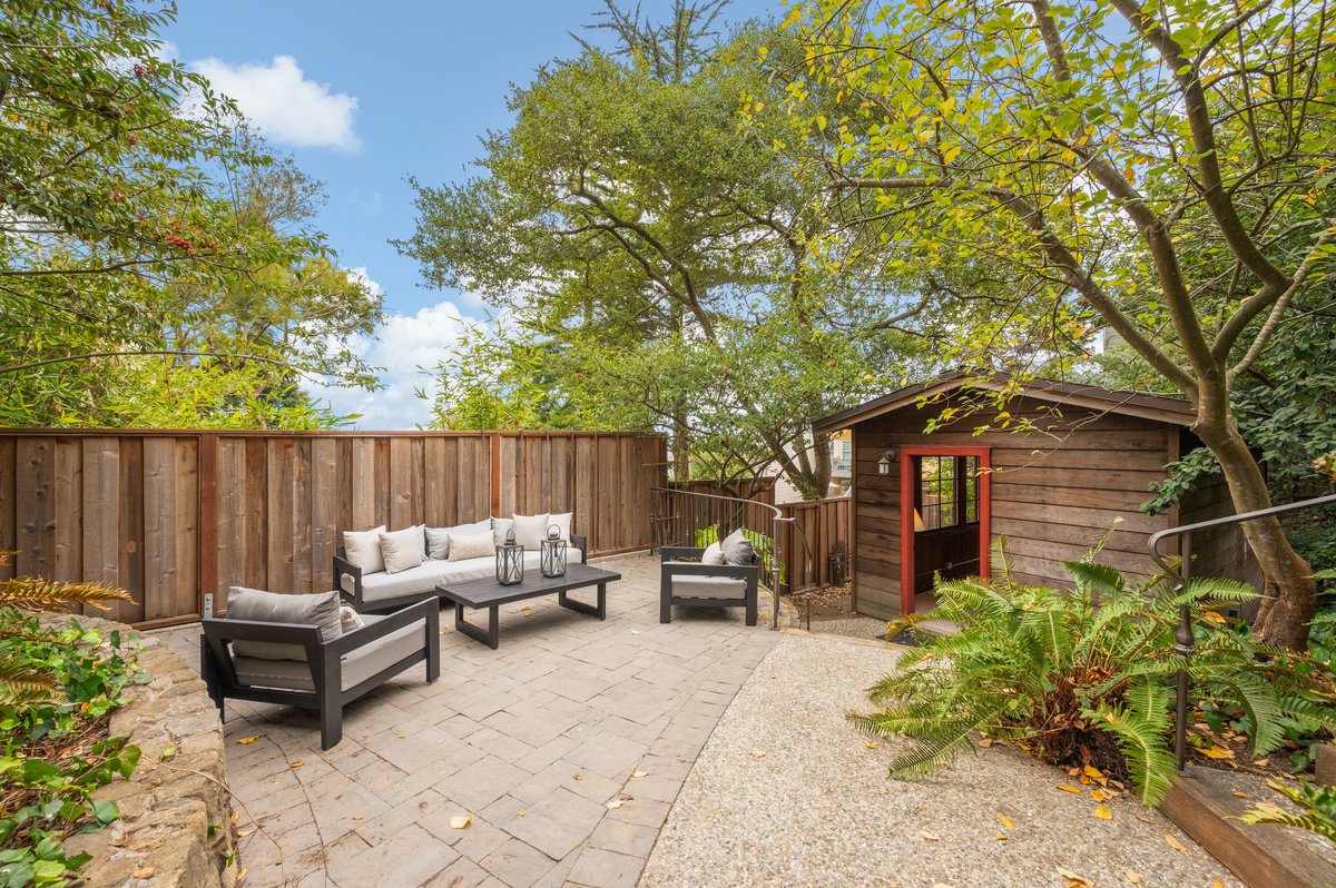 Outdoor patio area with cushioned seating, a small table, and lanterns on a stone path, surrounded by trees, ferns, and a wooden fence, with a small wooden structure in the background. Outdoor patio area with cushioned seating, a small table, and lanterns on a stone path, surrounded by trees, ferns, and a wooden fence, with a small wooden structure in the background.