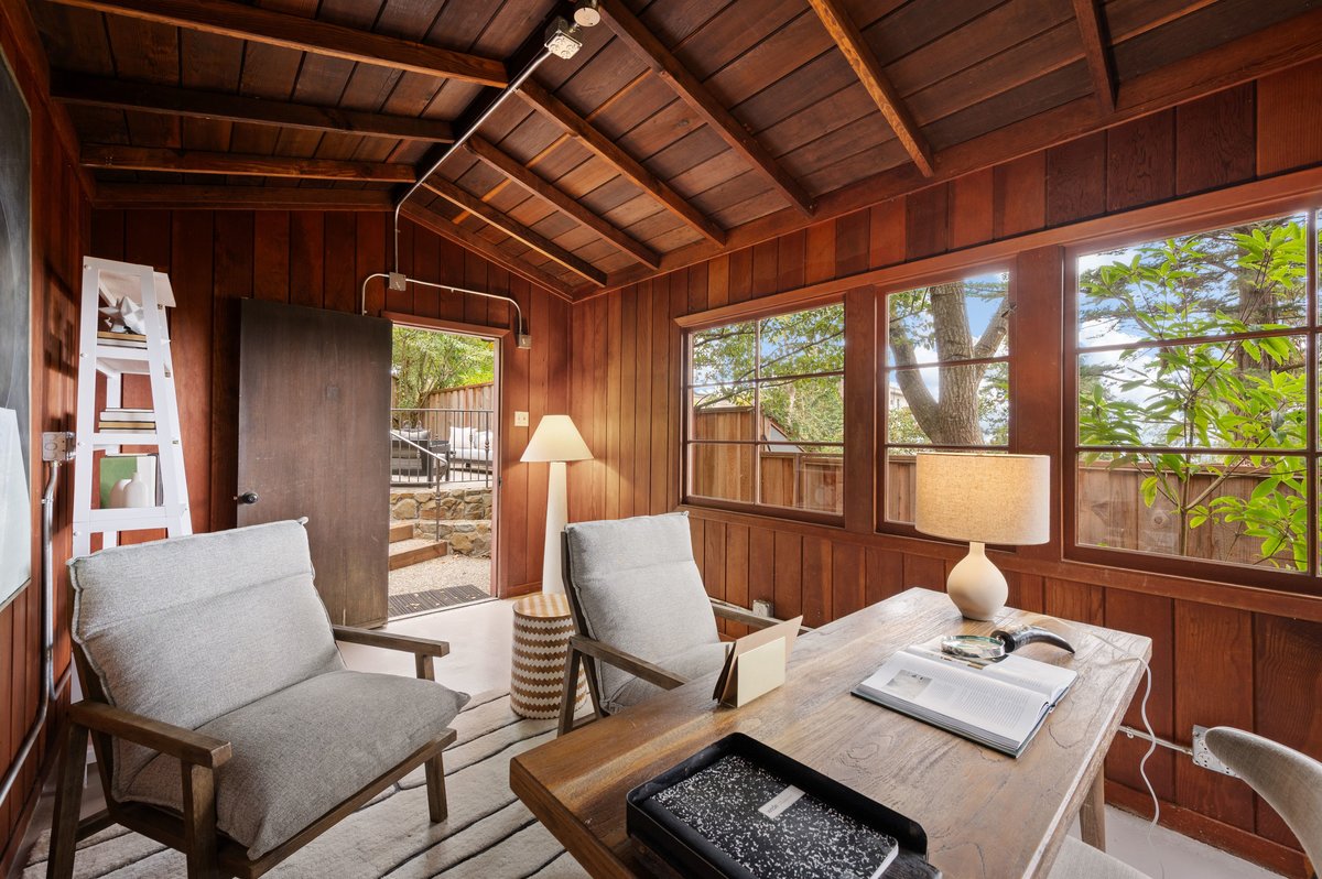 Wood-paneled home office with two cushioned chairs, a desk, a lamp, and large windows showing trees and a wooden fence outside. Wood-paneled home office with two cushioned chairs, a desk, a lamp, and large windows showing trees and a wooden fence outside.