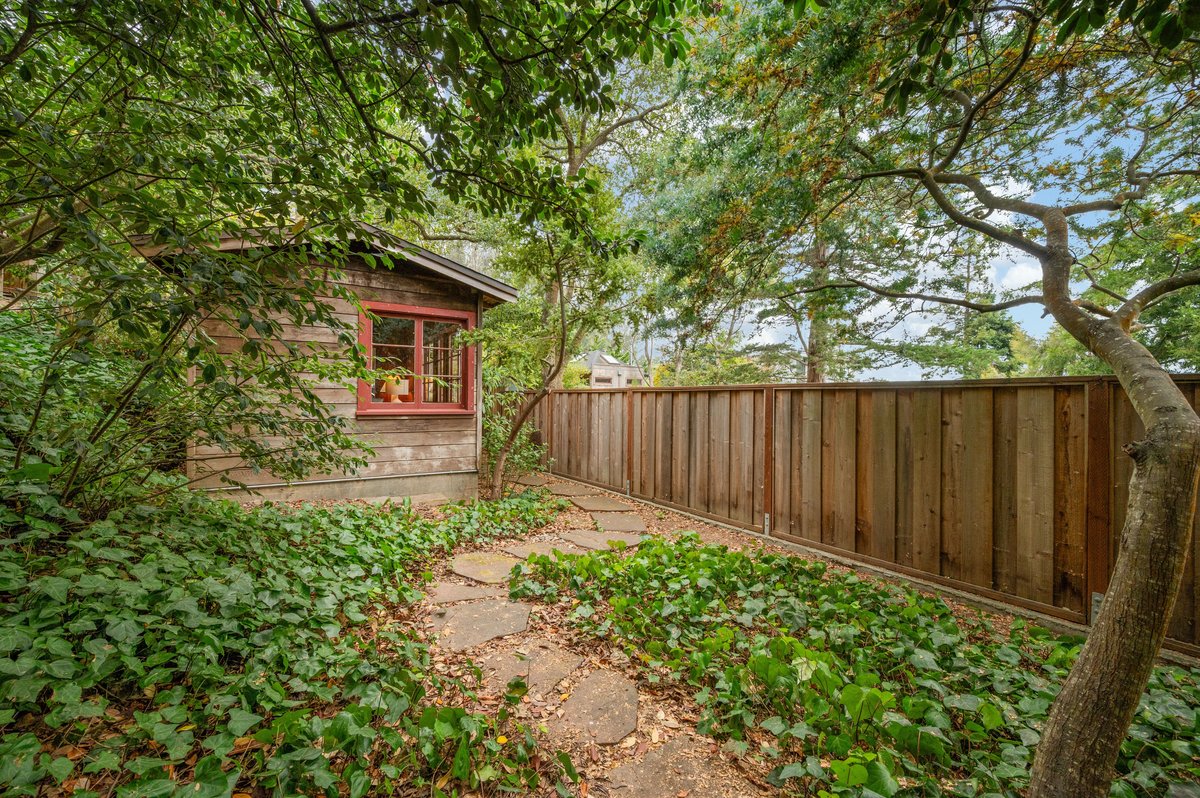 A small wooden structure with a window sits in a leafy backyard, featuring a stone path and a tall wooden fence under trees. A small wooden structure with a window sits in a leafy backyard, featuring a stone path and a tall wooden fence under trees.