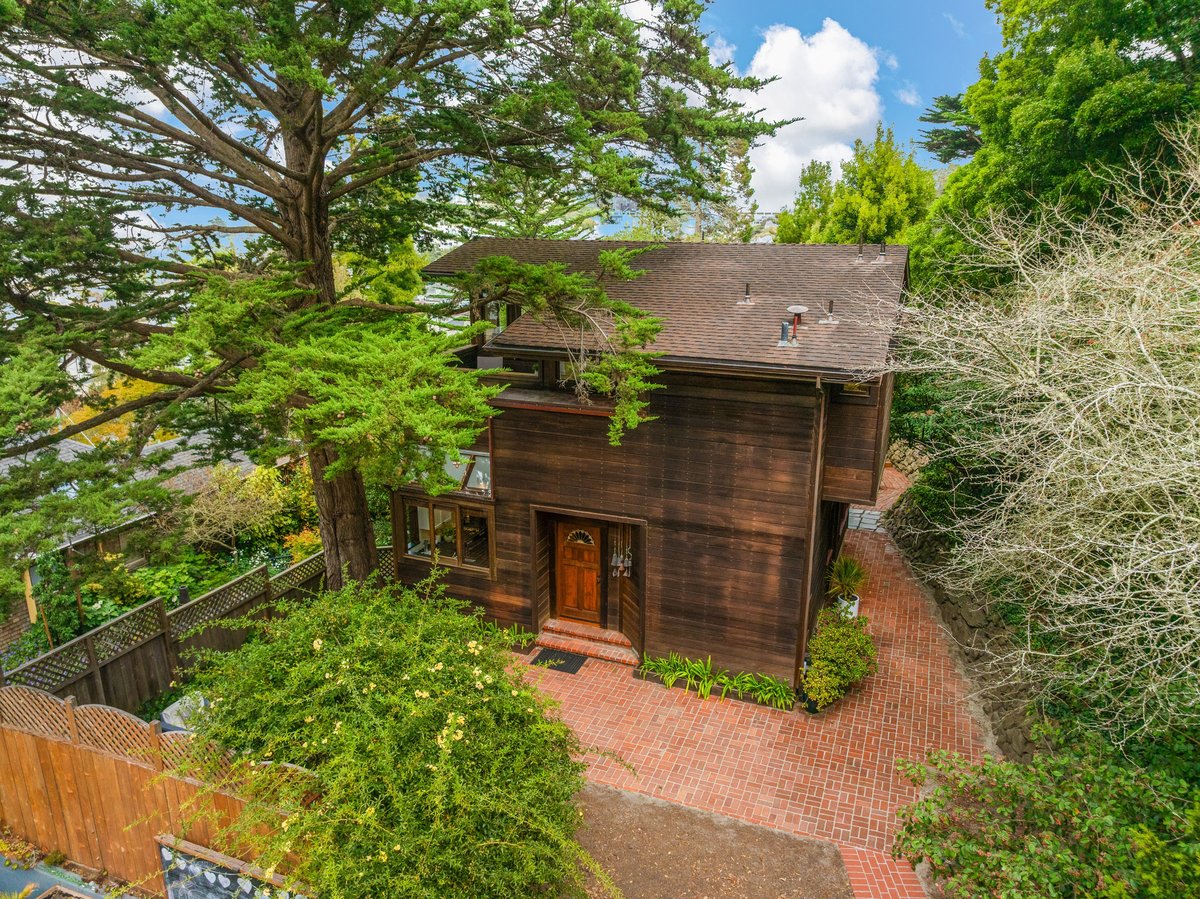 Two-story wooden house with a brick driveway, surrounded by tall trees and greenery, under a partly cloudy sky. Two-story wooden house with a brick driveway, surrounded by tall trees and greenery, under a partly cloudy sky.