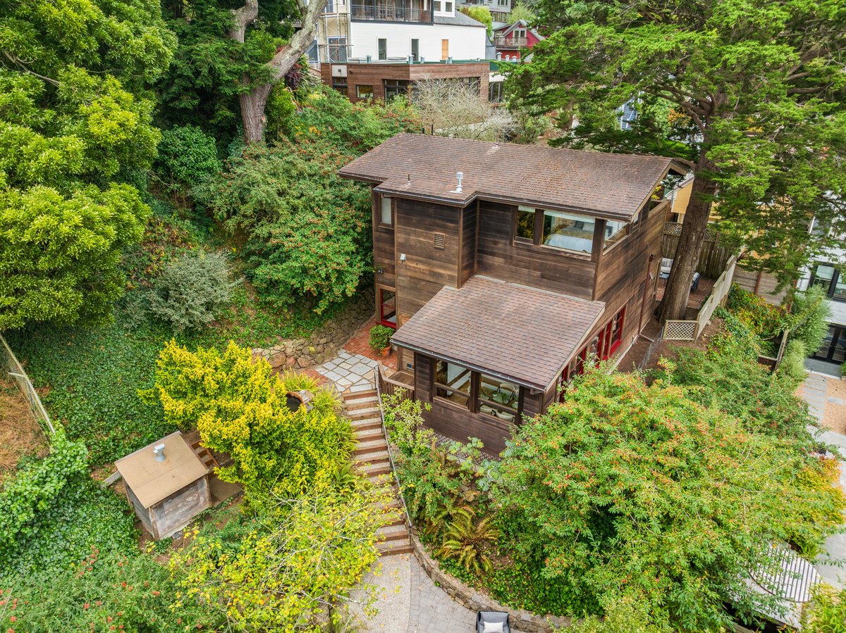 A two-story wooden house surrounded by dense greenery and trees, with a stone path and stairs leading to the entrance. A two-story wooden house surrounded by dense greenery and trees, with a stone path and stairs leading to the entrance.