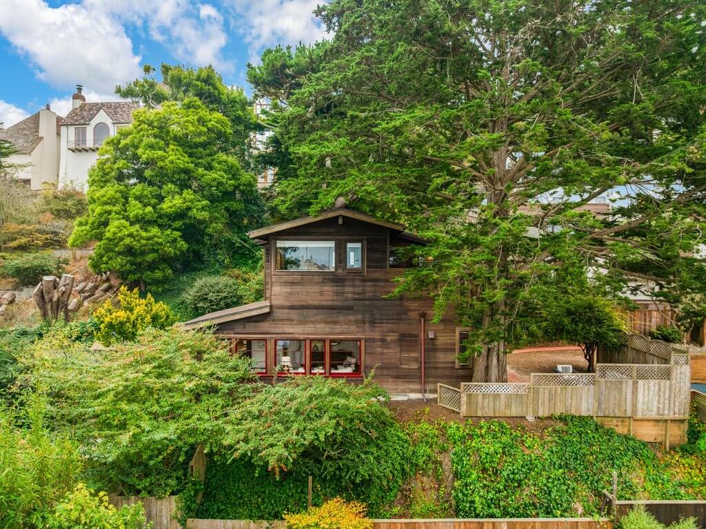 Two-story wooden house surrounded by dense green trees and shrubs, with a small wooden fence in front, under a partly cloudy sky.