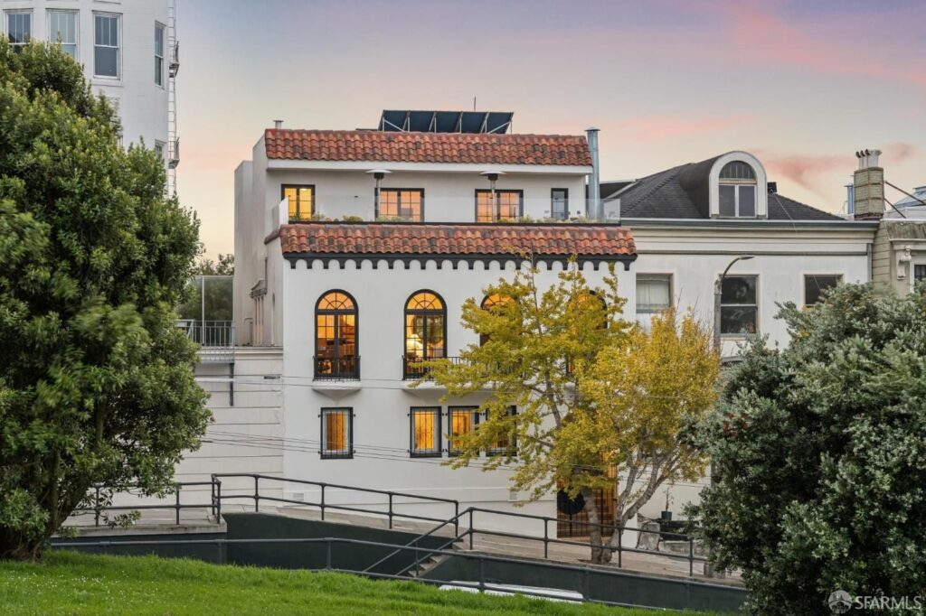 A white Mediterranean-style home with a red-tiled roof and arched windows, viewed from a park.