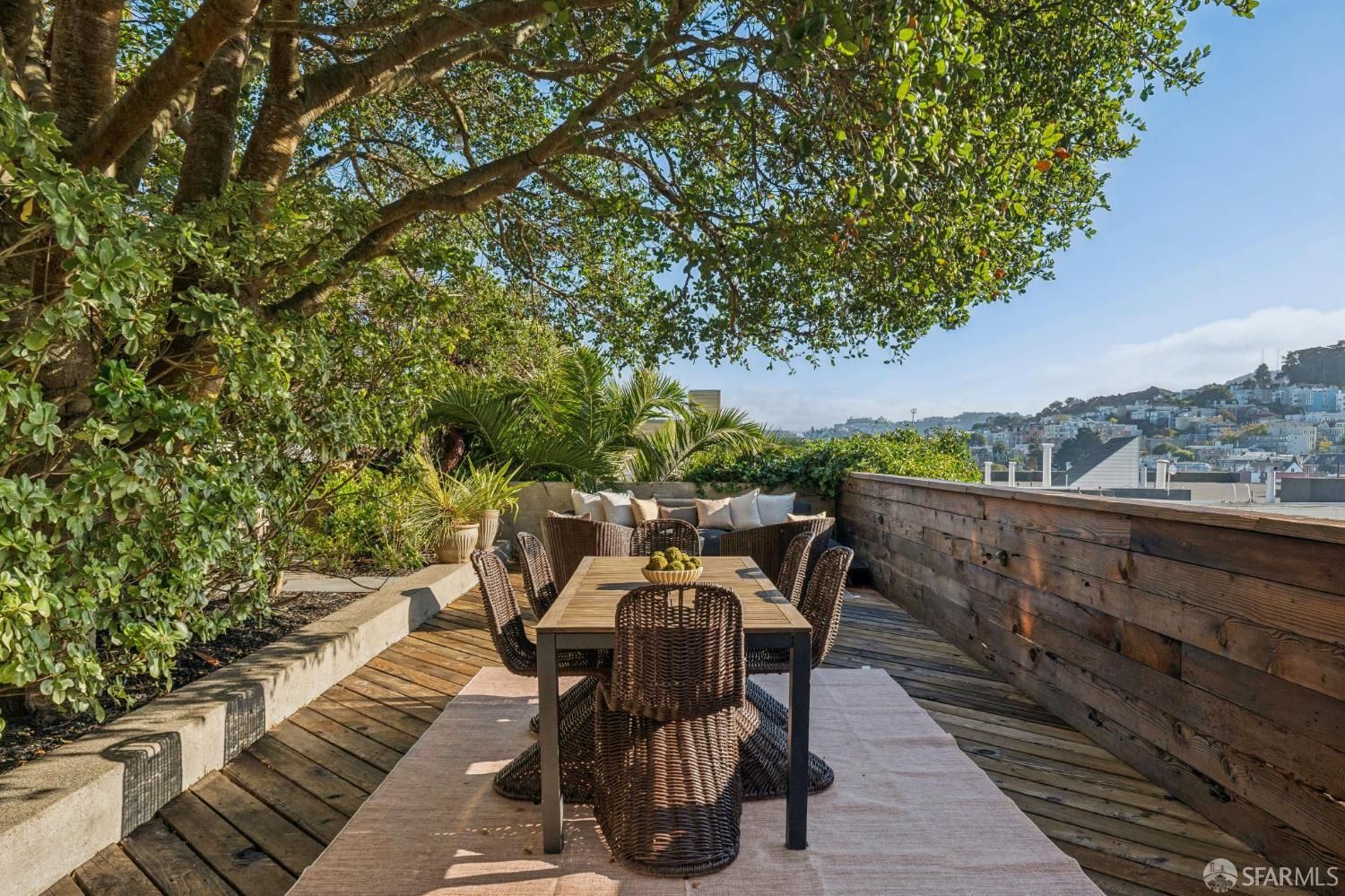 A wooden deck patio with a dining table, wicker chairs, and outdoor seating under a large tree, overlooking a hillside city. A wooden deck patio with a dining table, wicker chairs, and outdoor seating under a large tree, overlooking a hillside city.